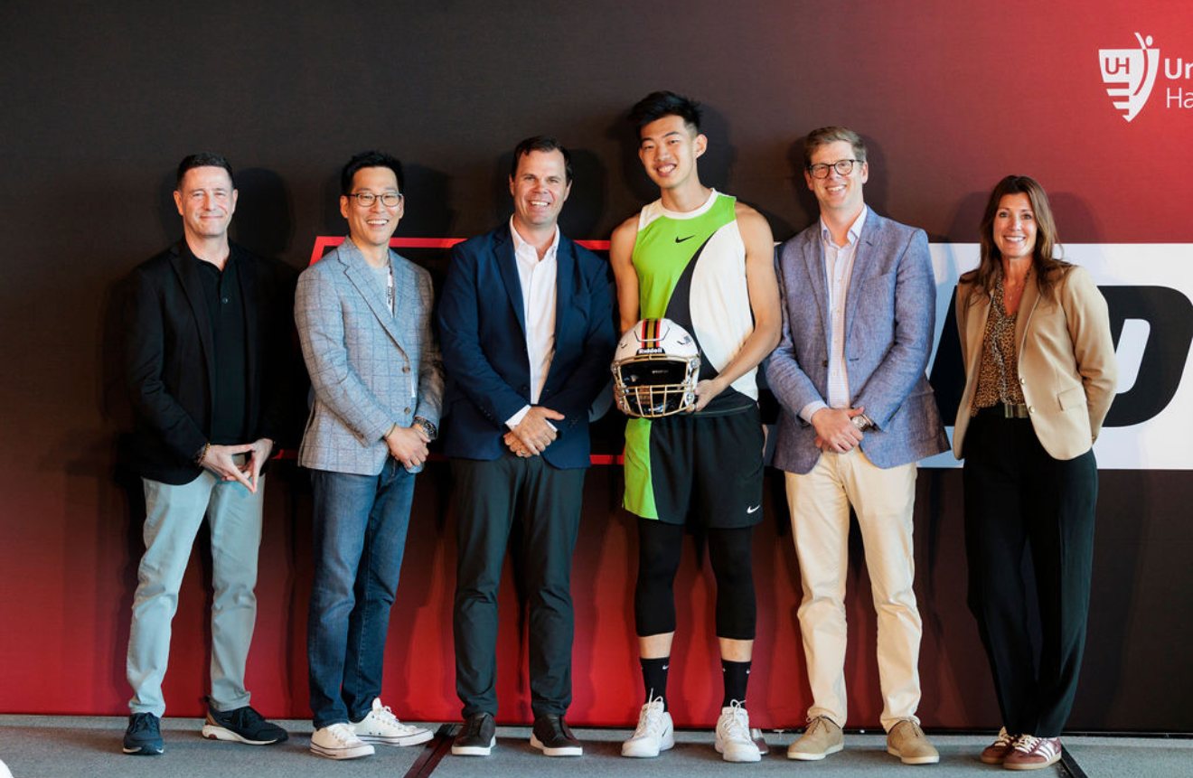 Hippos founder on stage with judges at the SportsLand Summit, wearing the Hippos soft exoskeleton and holding a Riddell helmet, University Hospitals branding behind.